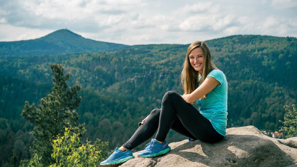 Smiling woman in blue sneakers sitting on a rock with mountain landscape in Jonsdorf, Germany.